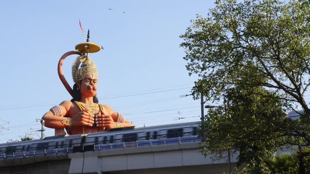 Locked-on shot of metro train moving in front of giant statue of lord Hanuman, Karol Bagh, New Delhi, India 