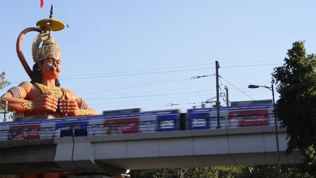 Locked-on shot of metro train moving in front of giant statue of lord Hanuman, Karol Bagh, New Delhi, India 