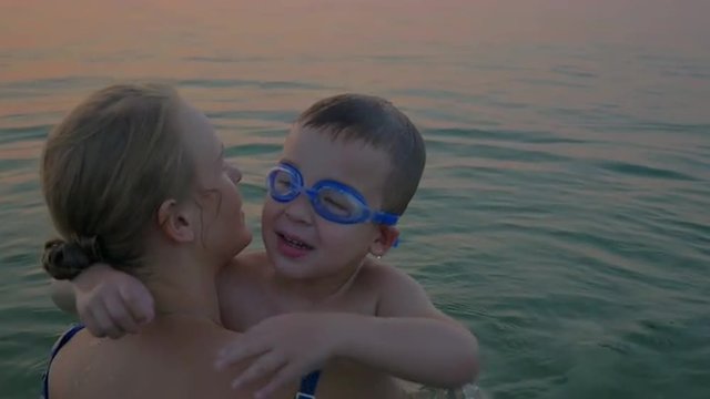 Slow motion of a boy in goggles embracing dear mother while they bathing in the sea. Mom kissing son and they looking at water. Happy family vacation