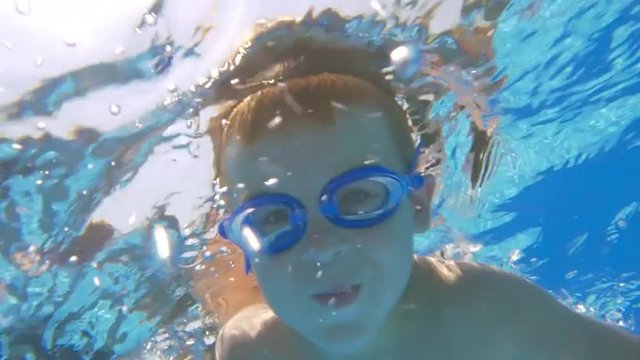 Slow motion of a little boy in goggles diving and swimming underwater in outdoor pool , then he coming up with following view of bright sunshine in clear blue sky
