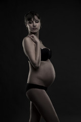 Young caucasian pregnant woman in black lingerie on gray studio background. Low key pregnancy concept.