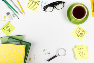 Office table desk with green supplies, blank note pad, cup, pen, glasses, crumpled paper, magnifying glass, flower on white background. Top view