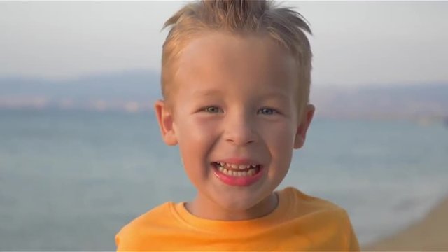 Close-up shot of a little boy in orange t-shirt with mohawk on the head looking to the camera and smiling. Happy careless child against sea background