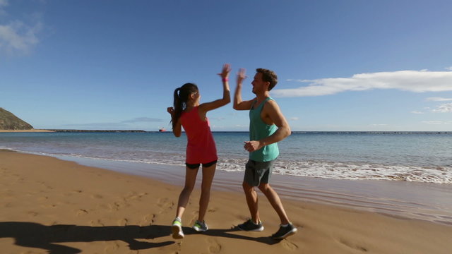 Running happy couple on beach giving high five cheering and celebrating, excited and joyful in celebration. Fit athletes, Asian woman and Caucasian man having fun on beach together.