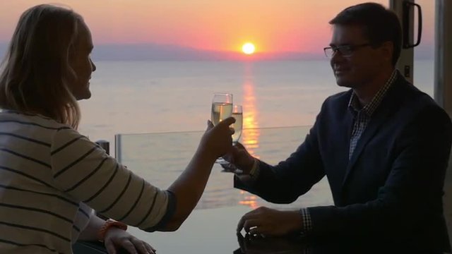Slow motion of young man and woman spending romantic evening. They clanging glasses and drinking wine at the balcony while observing scenic view of golden sunset over sea