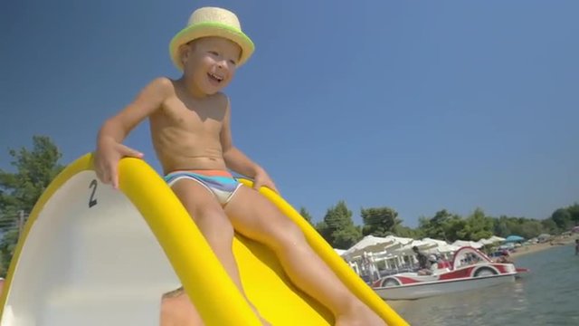 Slow motion of a happy little boy in summer hat riding on slider in the sea and father catching him in water. Jolly day on the beach 