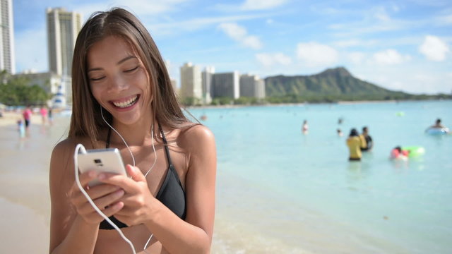 Woman using smartphone with earphones laughing on beach listening to music. Girl in bikini using mobile cell smart phone happy. Beautiful Asian Caucasian female model on Waikiki, Oahu, Hawaii, USA
