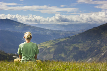 Naklejka na ściany i meble blonde girl sitting on grass and looking to mountains, meditating