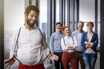 Corporate portrait of young black hipster businessman stretching braces, with his colleagues in background. Post processed with vintage film and sun filter.