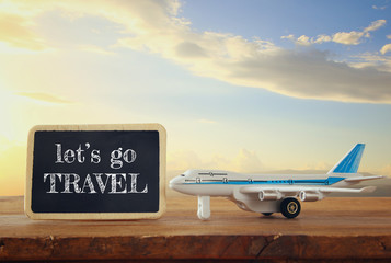 close up photo of toy airplane next to blackboard with text: LET'S GO TRAVEL, against sky with clouds. vintage filtered image, selective focus
