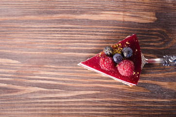 Piece of delicious raspberry cake with fresh raspberries, blueberry, currants and pistachios on shovel, wooden background. Free space for your text.