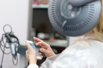 A blonde woman surfing the internet with her smartphone while having her hair coloring. Selective focus