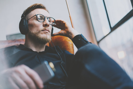 Portrait handsome bearded man glasses,headphones listening to music modern loft studio.Man sitting in vintage chair,holding smartphone and relaxing.Panoramic windows background.Horizontal, film effect