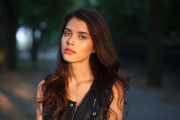 Closeup portrait of happy young beautiful brunette woman in black leather jacket posing on sunset outdoors  with blurry foliage background