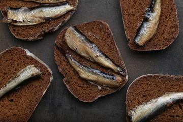 Snacks with sardines on black rye sliced bread. Riga traditional delicious dish. Smoked sprats on dark background.