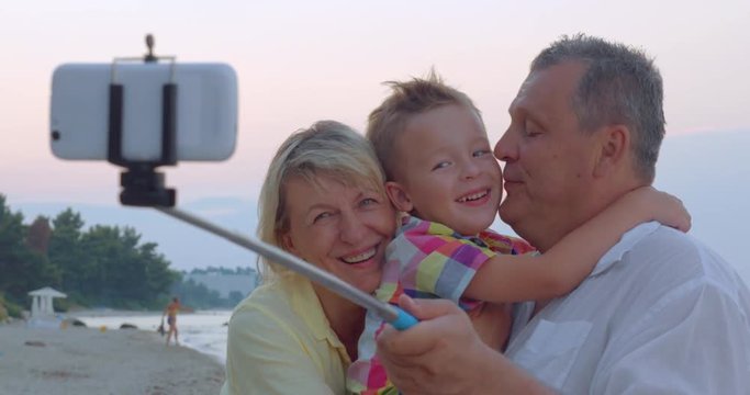 Cheerful little grandson and grandparents making selfie picture with mobile phone and monopod on the coast. They exchanging kisses to express love