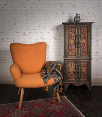 Living room corner including vintage orange armchair, ornamented cupboard and ornate scarf on a red carpet, dark wooden floor and white bricks wall