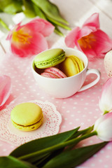 Fototapeta na wymiar A beautiful flowers pink tulips with colorful macaroons laid in cup on white wooden background with pink Lacy napkin
