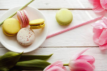 A beautiful flowers pink tulips with colorful macaroons laid on a white platter on white wooden background