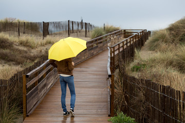 Une jeune femme sous son parapluie jaune sur un chemin de plage en bois 