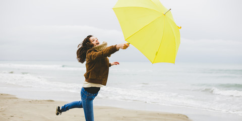 Fototapeta na wymiar Une jeune femme court , emportée par la force du vent avec son parapluie jaune sur une plage