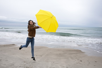 Une jeune femme court , emportée par la force du vent avec son parapluie jaune au bord des vagues