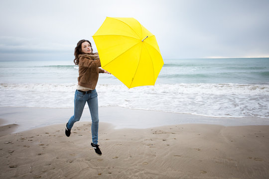 Une jeune femme emportée par la force du vent avec son parapluie jaune sur une plage