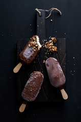 Chocolate dipped popsicles with chipped nuts on dark wooden board over black background