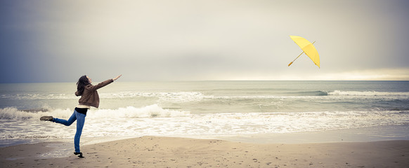 Une jeune femme court après son parapluie jaune envolé, sur une plage , avec teintes polaroid