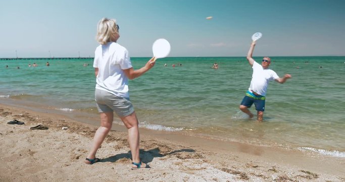 Mature couple is playing racket ball on the beach in sunny day, woman is standing on the sand, man is in water.