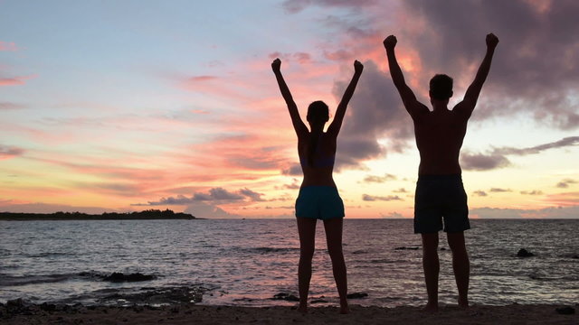 Success, achievement and accomplishment concept with fitness people cheering and celebrating of joy with arms raised outstretched up by ocean beach outside. Fitness couple having fun at sunset.