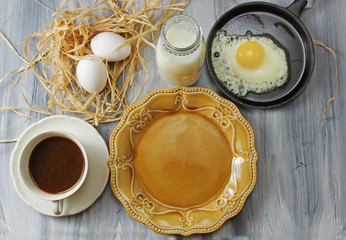 Overhead view of a table set with a plate, eggs, milk in a bottle, black coffee and fried egg in a pan on a grey textured background.