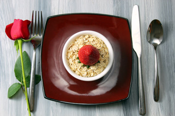 A romantic table setting with a Rose bud and a bowl of oats with strawberry on a rustic wooden textured background.