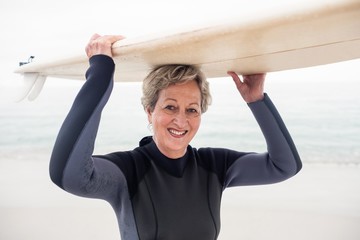 Portrait of senior woman in wetsuit carrying surfboard over head