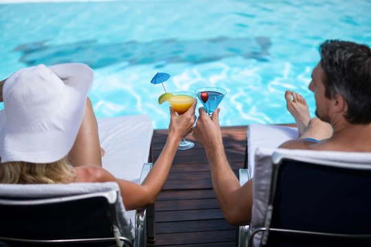 Rear View Of Couple Toasting Martini Glass While Relaxing On Sun Lounger Near Pool