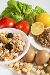Takeaway tuna salad served in a white plastic bowl next to a white plate with tuna chunks, lemon, olives, croutons, and fresh eggs, cucumbers, tomatoes and lettuce.