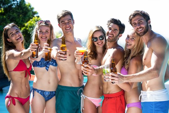 Group of happy friends showing beer bottles and glass of cocktail near pool
