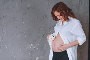 Pregnant woman in a white man's shirt, a studio portrait. Looking at the midsection. With a Teddy bear in front of him.