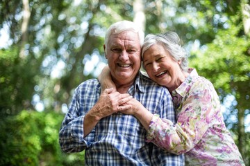 Happy senior woman embracing from behind husband against trees
