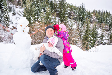 Obraz premium Happy father and his two daughters building a snowman on winter day. Family playing on nature with forest in background. Christmas time