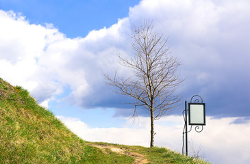 hill, turning path, bare tree, sky with clouds and empty small decorated billboard with free space for your text during beatiful sunny spring day