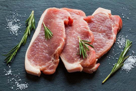 Raw pork steaks with rosemary on black stone background.