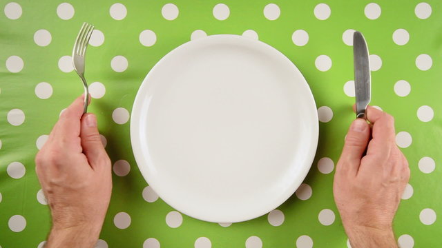 Top view of nervous hungry male hands at dinner table holding fork and a knife above empty flat white plate, dieting concept.