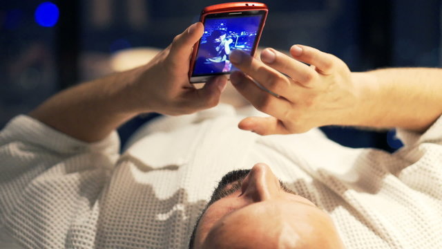Man browsing photos on smartphone lying on bed at home at night
