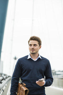 Germany, Hesse, Frankfurt, portrait of young businessman walking on a bridge  with briefcase and smartphone