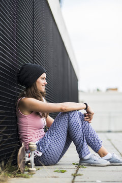 Smiling teenage girl with skateboard sitting in front of a black facade