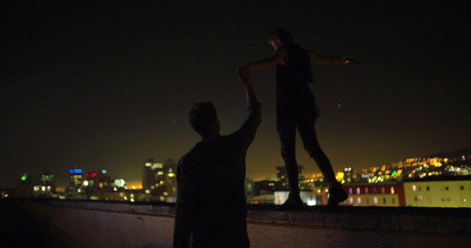 Man is holding hand with girlfriend who is balancing on rooftop edge
