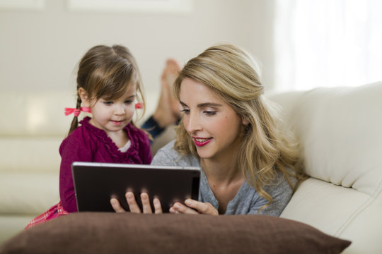 Mother and her little daughter together on couch in the living room looking at digital tablet