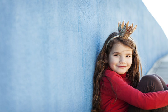 Portrait of smiling little girl with a crown leaning against blue wall