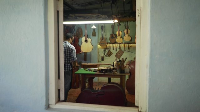 Lute maker shop and classical music instruments: young adult artisan opening his shop. He fixes an old classic guitar lacking a string. Dolly shot